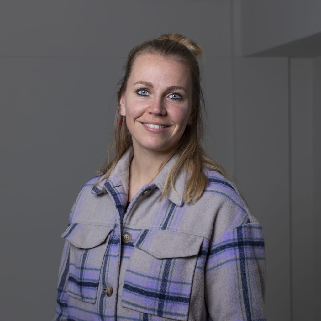 Vrouw met blond haar en blauwe ogen in een geruite jas, neutrale achtergrond – tevreden cliënt van Myrthe, bedrijfspsycholoog